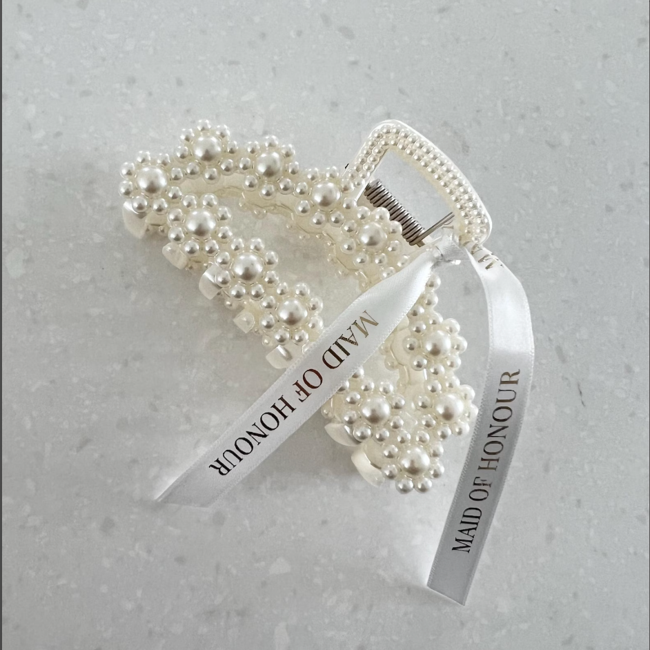 Two pearl-bead hair claw clips with white ribbon tags that read 'MAID OF HONOUR', photographed on a light speckled stone surface.