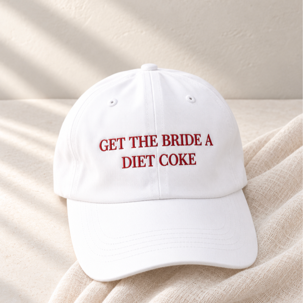 A white baseball cap with red embroidered text reading 'GET THE BRIDE A DIET COKE' resting on a beige textured fabric surface with soft shadow lighting.