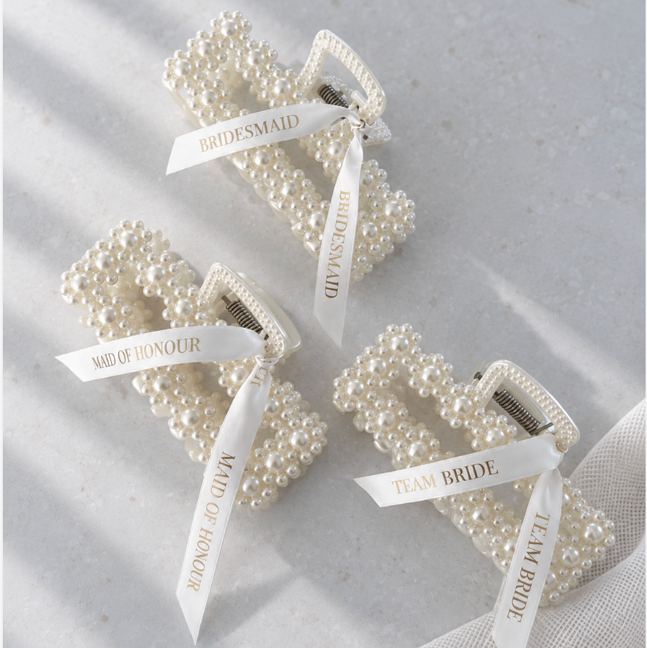 Three pearl-beaded hair claw clips arranged on a light fabric surface. Each clip is tied with a white ribbon printed with bridal-themed phrases: 'BRIDESMAID,' 'MAD OF HONOUR,' and 'TEAM BRIDE.