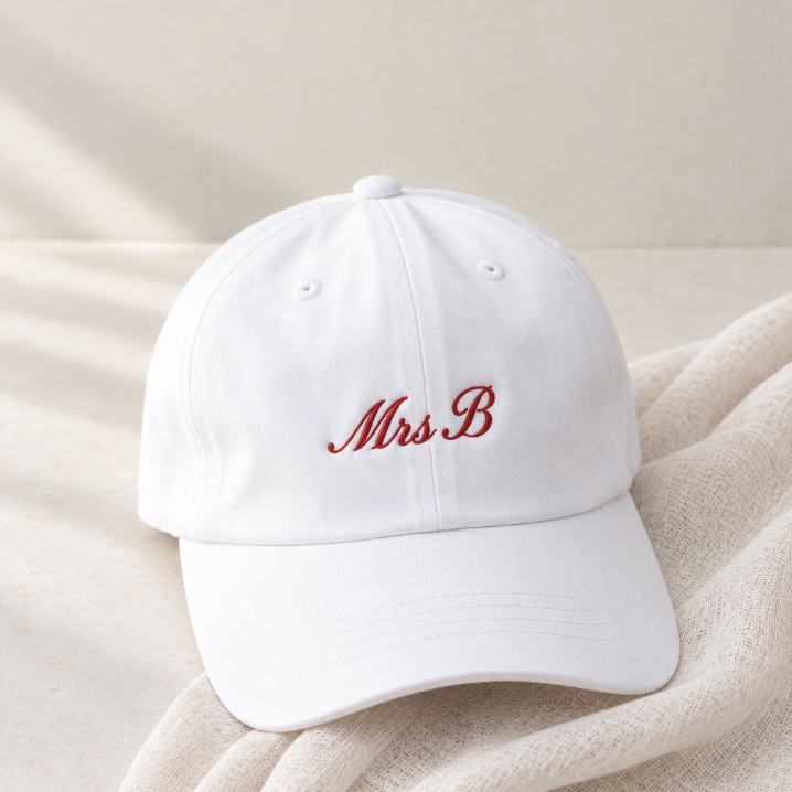 A white baseball cap with 'Mrs. B' embroidered in red cursive on the front, resting on a textured beige fabric against a neutral beige background.