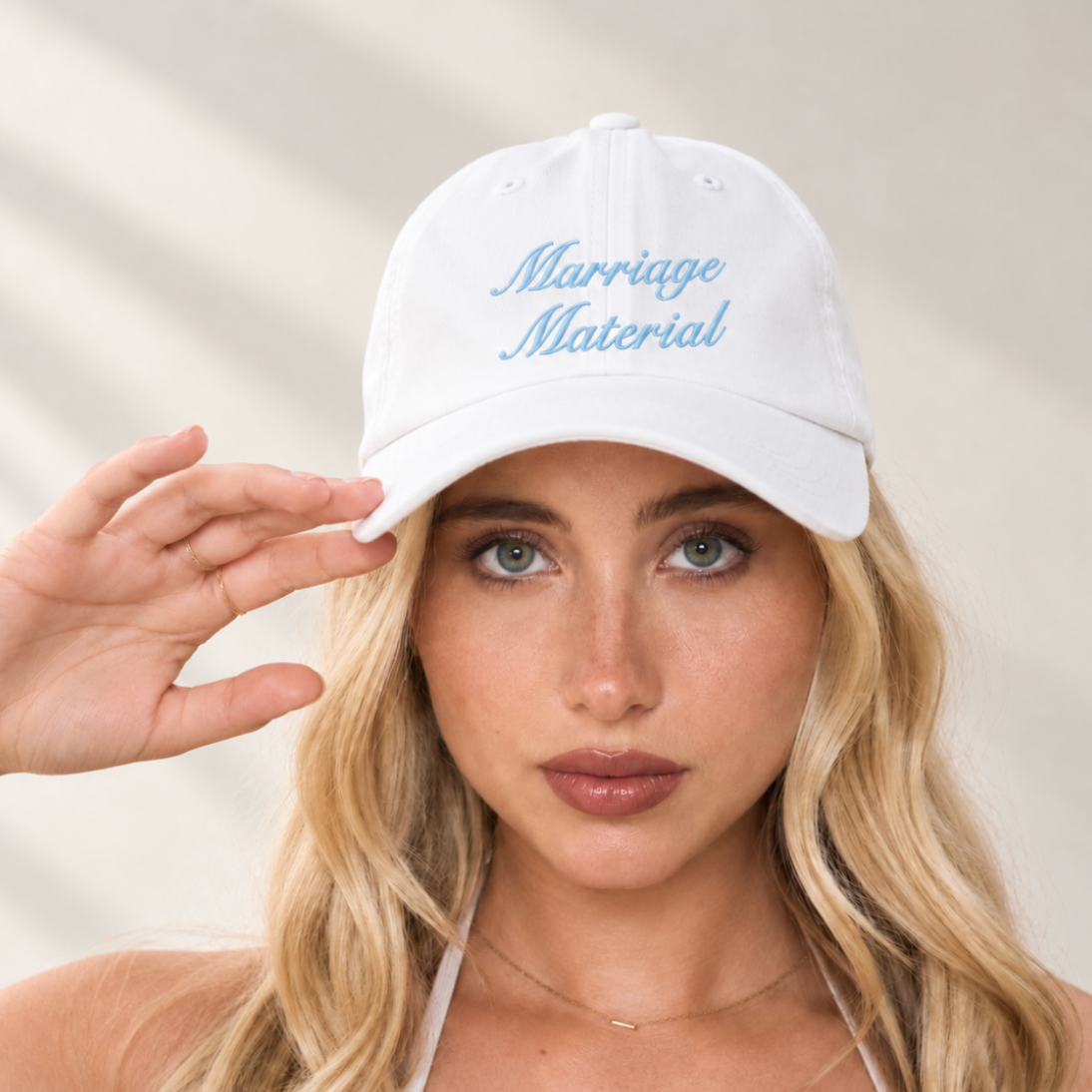 A blonde woman wearing a white baseball cap with 'Marriage Material' embroidered in light blue script on the front, posing with her hand near her face against a softly lit neutral background.