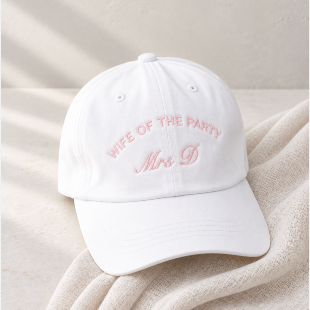 A white baseball cap with pink embroidered text reading 'WIFE OF THE PARTY Mrs D' resting on a soft beige textured blanket.
