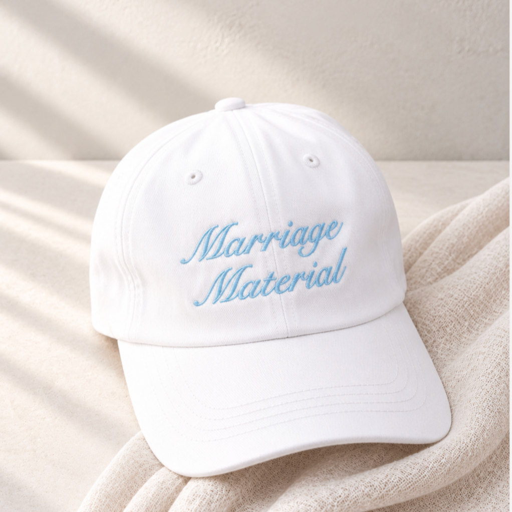A white baseball cap with light blue embroidered text reading 'Marriage Material' resting on a beige textured blanket with soft window shadows.