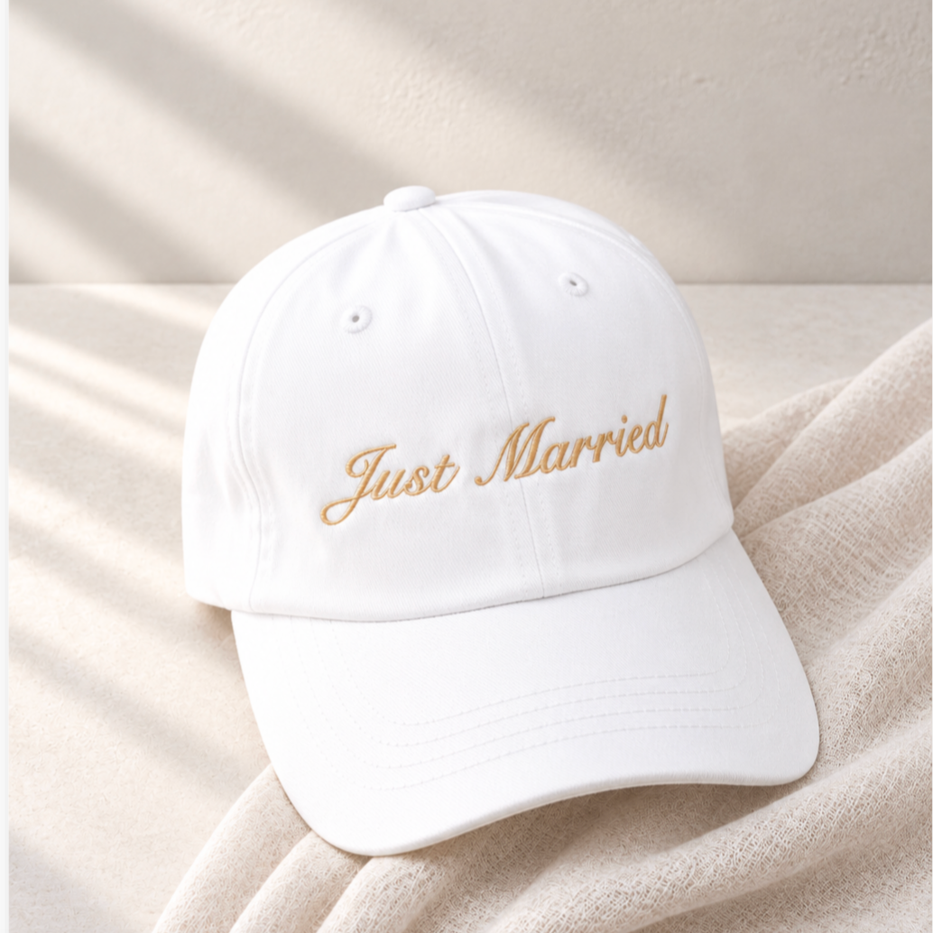 A white baseball cap with 'Just Married' embroidered in gold script on the front, resting on a soft beige textured blanket with dappled sunlight and shadows.