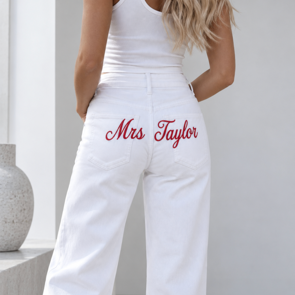 A woman shown from the back wearing white wide-leg jeans with 'Mrs Taylor' embroidered in red script on the back pocket, paired with a white tank top in a bright indoor setting.