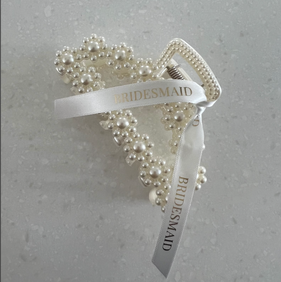A pearl-embellished bridal hair clip with a white satin ribbon printed with the word 'BRIDESMAID' in gold, photographed on a light gray textured surface with water droplets.