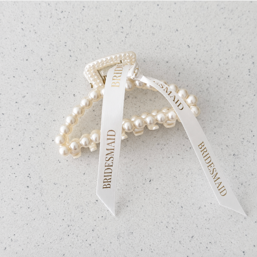 Three pearl-bead bridal bracelets with white ribbon tags printed with the word 'BRIDESMAID', arranged on a light speckled stone surface.