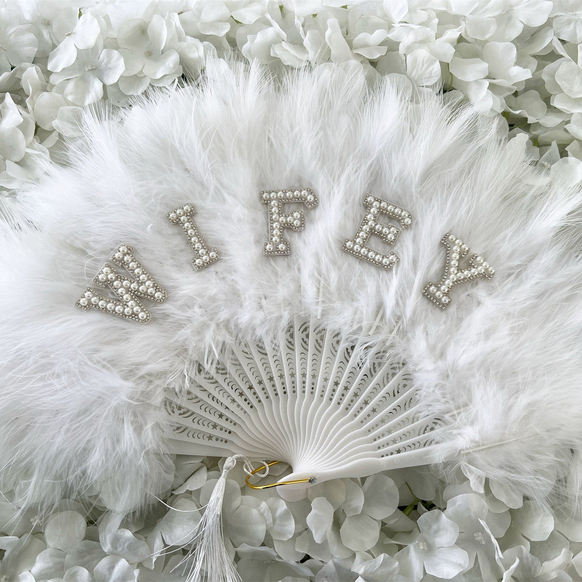 A white feather fan with pearl-embellished lettering spelling 'WIFEY' resting on a bed of white flower petals.