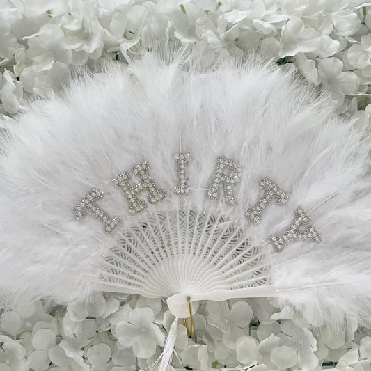 A white feather fan with rhinestone lettering spelling 'THIRTY', displayed against a background of white flowers.