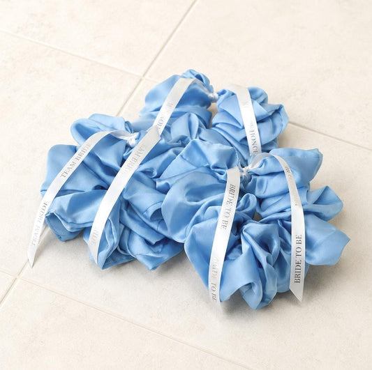 Three light blue satin hair scrunchies tied with white ribbons printed with 'BRIDE TO BE', arranged on a light beige tiled floor.